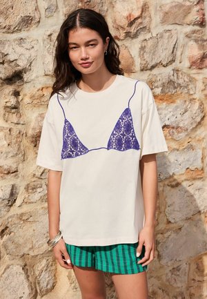 Fiatal nő sötét hullámos hajjal fehér pólót visel lila csipke melltartó mintával és zöld csíkos rövidnadrággal, egy kőfal előtt áll.