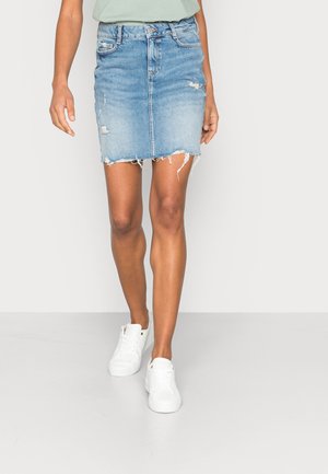 Vrouw draagt een lichtblauwe versleten spijkerrok met rafelige zoom en witte sneakers, staand tegen een effen lichte achtergrond.