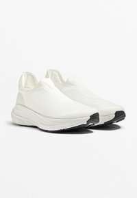 Biele tenisky na slip-on s textúrovaným pleteným zvrškom, elegantným dizajnom a čiernou gumovou podrážkou. Bez viditeľných šnurovacích systémov a s okrúhlym tvarom špičky.