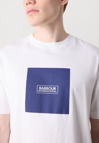 Bílý bavlněný tričko s kulatým výstřihem, na přední straně s modrým obdélníkovým logem. Text zní "BARBOUR INTERNATIONAL" v bílé barvě.