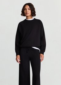 Sweatshirt noir à coupe décontractée avec des manches raglan, associé à un pantalon noir à jambes larges avec une taille à cordon de serrage et un sous-shirt blanc.
