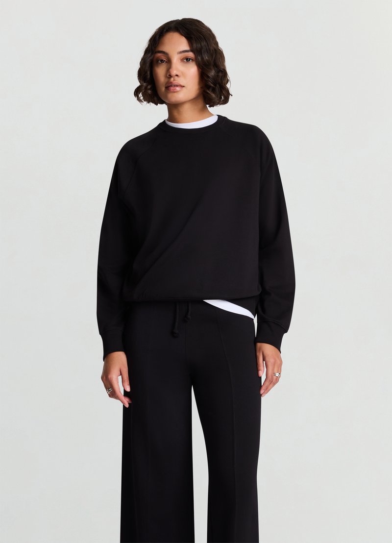 Sweatshirt noir à coupe décontractée avec des manches raglan, associé à un pantalon noir à jambes larges avec une taille à cordon de serrage et un sous-shirt blanc.