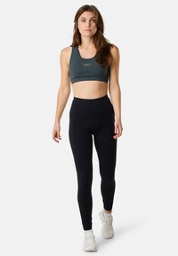 Grön sportbehå och höga svarta leggings i slät, elastisk tyg. Behån har rund hals och diskret logotypdetalj.