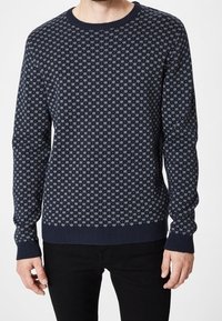 Mörkblå rundhalssweater med ett ljusblått snöflinge-mönster, bärs tillsammans med svarta byxor, sedd från halsen till höfterna.