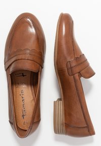 Une paire de mocassins en cuir marron avec un détail de bride à bords festonnés, un petit talon empilé et un bout arrondi, montrée en vue de dessus et de côté.