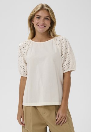 Uśmiechnięta blondynka w kremowej bluzce z bufkami i ażurowymi detalami oraz jasnobrązowych spodniach z wysokim stanem, stojąca na tle jasnoszarego tła.