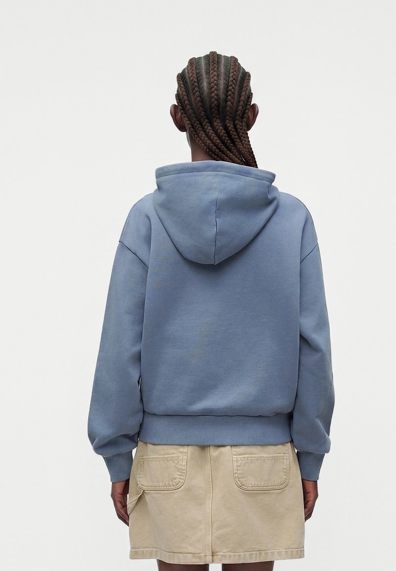 Person with braided hair wearing a blue hoodie and beige skirt stands facing away against a plain light background.