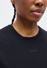 Κοντινό πλάνο ατόμου που φοράει μαύρο μπλουζάκι με στρογγυλή λαιμόκοψη και πλεγμένα μαλλιά, δείχνοντας τη λέξη "BORG" τυπωμένη στο στήθος.