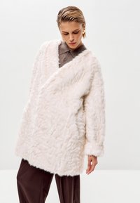 Abrigo de piel sintética blanca con una superficie texturada y esponjosa, diseño envolvente, mangas anchas y un aspecto suave, superpuesto a una camisa marrón con cuello.