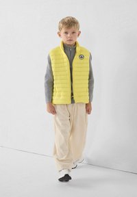 Gele doorgestikte bodywarmer met hoge kraag, gedragen over een grijze ritsvest, gecombineerd met crèmekleurige corduroybroeken en witte sportschoenen.