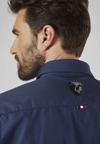 Chemise bleu marine avec un insigne représentant un coq doré et le texte "FRANCE". Comprend une étiquette tricolore contrastante à l'arrière. Texture lisse.