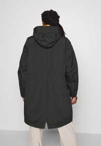 Parka noire imperméable avec capuche, taille ajustable et silhouette allongée. Caractérisée par un design minimaliste et une doublure intérieure contrastante.