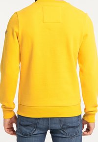 Felpa gialla con maniche lunghe, polsini e orlo a coste. Presenta una toppa quadrata sul retro. Abbinata a jeans aderenti blu scuro.