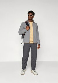 Pilka fliuso striukė, geltoni marškinėliai, pilkos kelnės ir balti sportbačiai. Aksesuarai: juoda kuprinė ir balti akiniai nuo saulės.