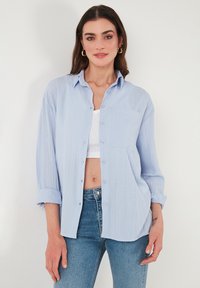Camicia azzurra chiara con un tessuto strutturato, maniche arrotolate e una singola tasca sul petto, abbinata a jeans aderenti.