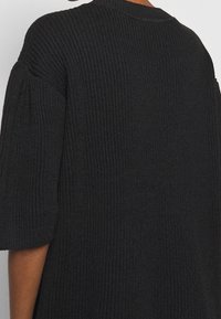 Pull en tricot noir avec un motif texturé, des manches larges et une coupe ample. Le tissu semble doux et confortable, idéal pour le superposition.
