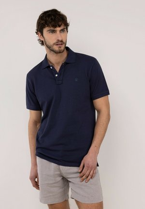 Hombre con cabello rizado oscuro lleva polo azul marino y pantalones cortos gris claro, de pie contra un fondo liso y claro mirando hacia un lado.