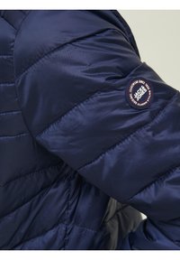 Námořnická modrá prošívaná bunda s prošívanou texturou, kulatou nášivkou s logem na rukávu a hladkým, lesklým povrchem.