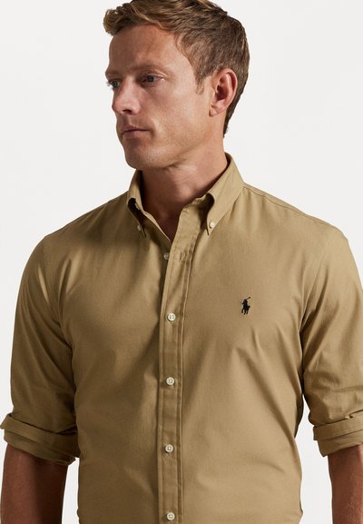 Beige skjorte med knapper laget av bomull, med slim fit, som har lange ermer rullet opp og en svart brodert logo på brystet.