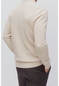 Homme portant un pull beige texturé et un pantalon marron foncé, une main dans la poche, vu de dos sur un fond uni.