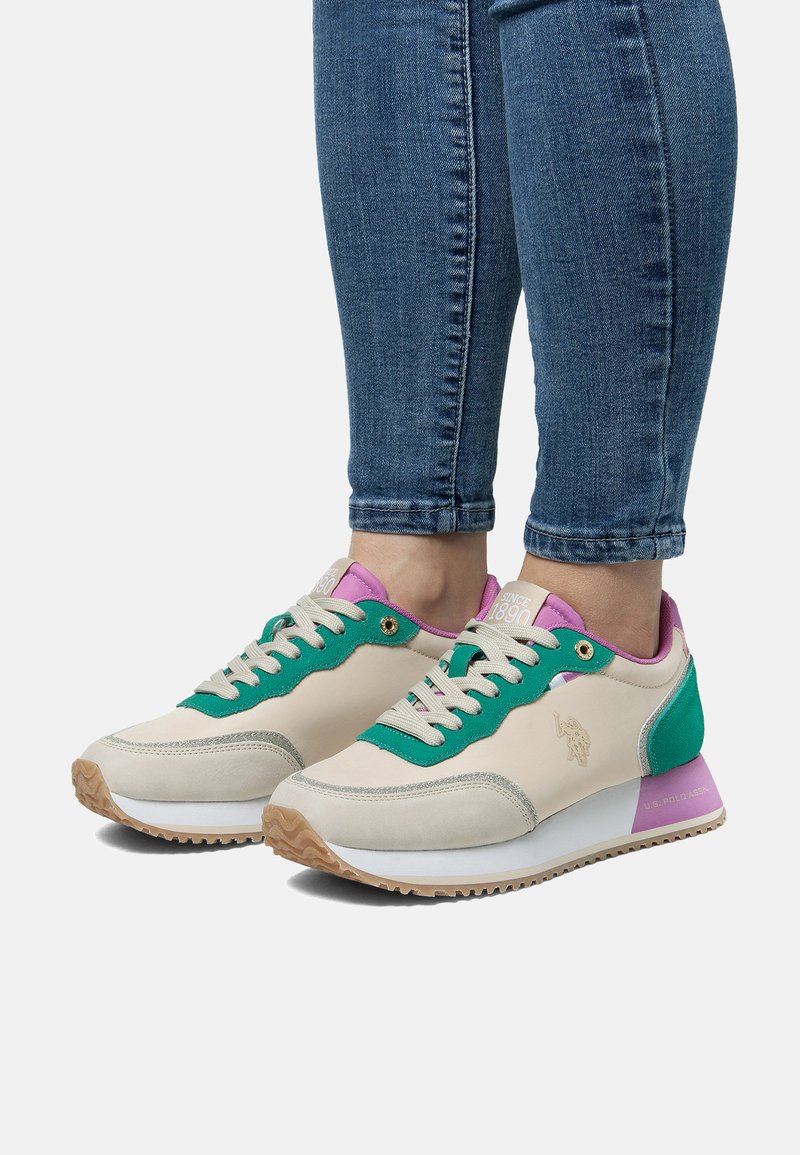 Piedi che indossano scarpe da ginnastica bianche, verdi e viola con suole beige e jeans slim blu su uno sfondo chiaro e semplice.
