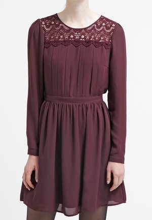 Langärmliges burgunderfarbenes Kleid mit Spitzenpasse, plissiertem Oberteil, tailliertem Bund und gerafftem Rock, mittellang bis zum Oberschenkel getragen.