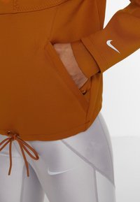 Pomarańczowa sportowa bluza z kapturem, przednią kieszenią, ściągaczem u dołu i prążkowanymi mankietami. Do tego błyszczące szare leginsy z białym logo.