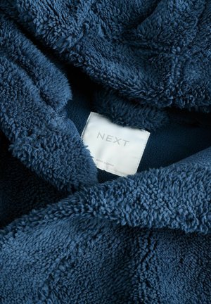Tissu en polaire bleu marine avec une surface douce et texturée. L'étiquette indique "NEXT" avec "FABRIQUÉ EN CHINE" en dessous. Design confortable et surdimensionné.