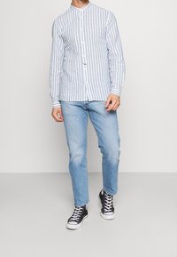 Homme portant une chemise à boutons rayée verticalement bleu clair et blanc, un jean bleu clair et des baskets montantes noires, debout devant un fond uni.