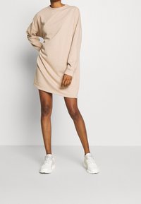 Robe sweat beige oversize avec manches longues, col rond et texture douce, associée à des baskets de sport blanches.