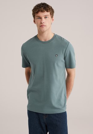 Jonge man met krullend haar, gekleed in een zeegroen T-shirt met korte mouwen en donkerblauwe spijkerbroek, staand tegen een effen beige achtergrond.