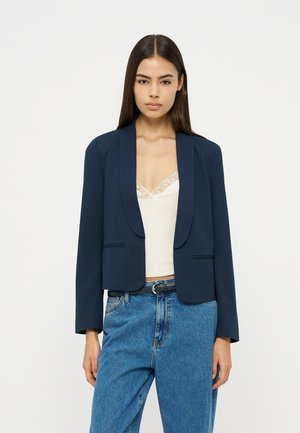 Γυναίκα με ίσια σκούρα μαλλιά, που φοράει μπλε σκούρο σακάκι πάνω από λευκό τοπ με δαντέλα και μπλε τζιν ψηλόμεσο, μπροστά σε απλό φόντο.