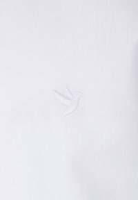 Tela de algodón blanca con un pequeño diseño de colibrí bordado en blanco. La textura parece suave y limpia.
