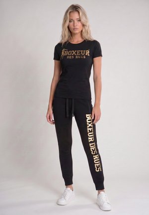 Jeune femme debout portant un t-shirt noir et un pantalon avec le texte doré « Boxeur Des Rues » et des baskets blanches sur un fond clair uni.