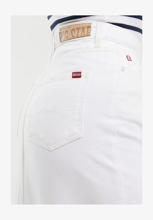 Baltie džinsi ar augstu vidukli, ar brūnu ādas etiķeti aizmugurē un mazu sarkanu logotipa birku aizmugurējā kabatā.