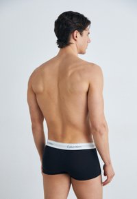 Crne boksačice s bijelim pojasom Calvin Klein, glatke teksture i uskog kroja, prikazane iz stražnjeg kuta.