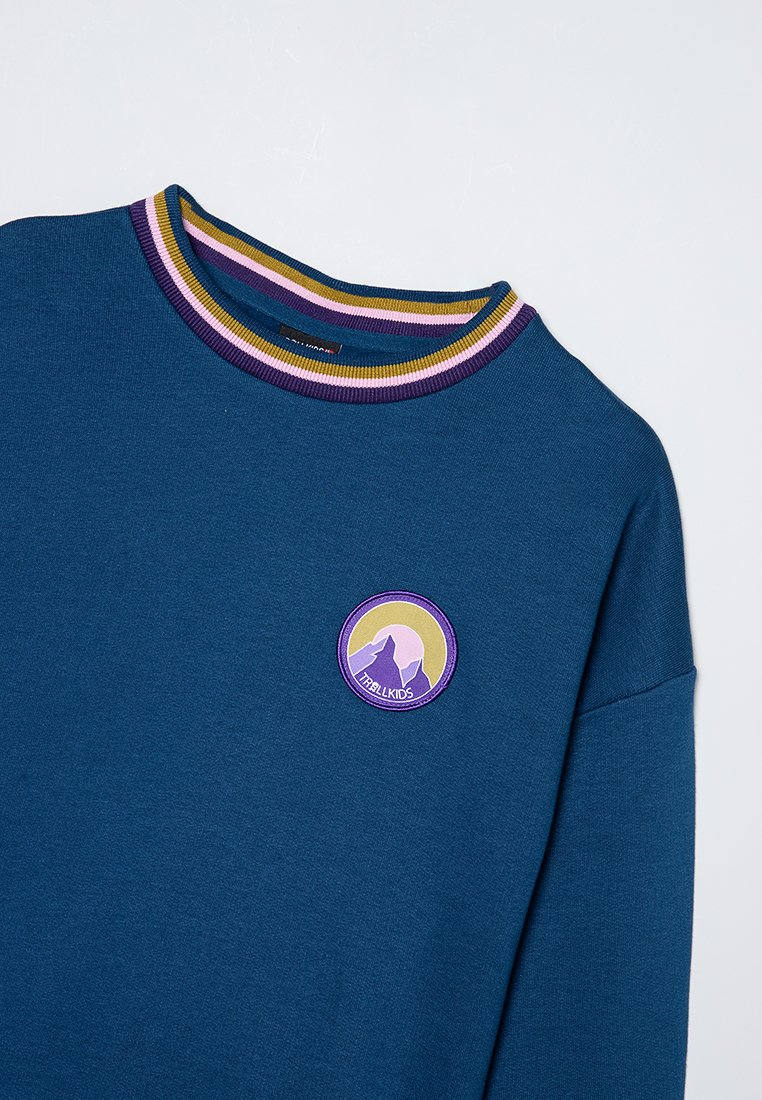 Blaues Sweatshirt mit einem runden lila Logo, das Berge zeigt. Gerippter Kragen in grünen, gelben und lila Streifen. Weicher Stoff, lockere Passform.