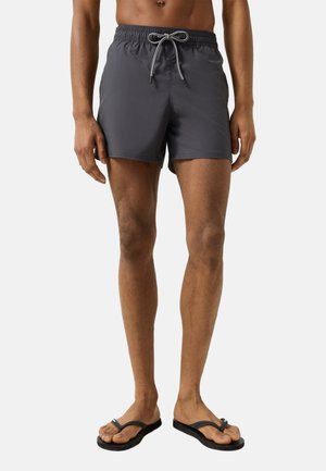 Man wearing dark gray swim shorts with a tied drawstring and black flip-flops, standing against a plain background.