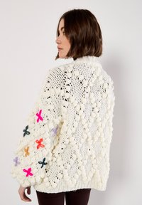 Cardigan spesso lavorato a maglia bianca con filato testurizzato, motivo a rete aperta e fiori colorati ricamati sulle maniche.