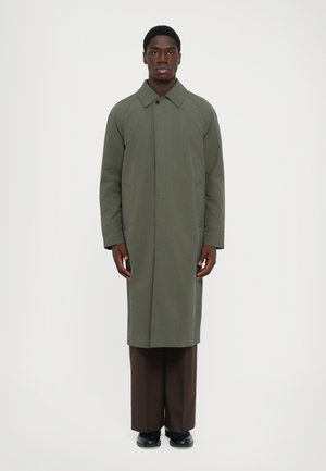 Man staat rechtop met zijn gezicht naar voren, gekleed in een lange olijfgroene jas, bruine wijde broek en zwarte schoenen, tegen een effen witte achtergrond.