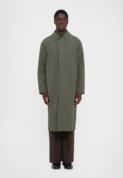 Man staat rechtop met zijn gezicht naar voren, gekleed in een lange olijfgroene jas, bruine wijde broek en zwarte schoenen, tegen een effen witte achtergrond.