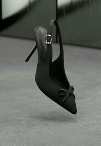 Svart spetsig slingback med hög klack och en liten rosett på framsidan samt ett guldspänne på sidoväskan, svävande ovanför en grå yta.