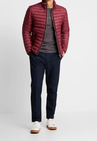 Bordeauxrote gesteppte Jacke mit hohem Kragen, graues T-Shirt darunter, marineblaue Hose und weiße Sneaker mit braunen Sohlen.