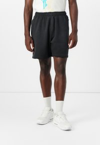 Črne športne kratke hlače z elastiko v pasu, z robom brez obdelave, obute z belimi supergami in nogavicami do gležnjev.