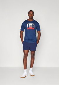 Tumesinine Under Armour t-särk punase ja valge graafilise logoga, kokku sobitatud tumesiniste lühikeste pükste ja valgete spordijalatsitega. Lihtne ja funktsionaalne disain.