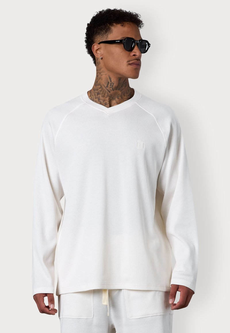Weißes Langarmshirt mit V-Ausschnitt, strukturiertem Stoff und Raglanärmel. Kleines Logo auf der Brust. Kombiniert mit cremefarbenen Jogginghosen.