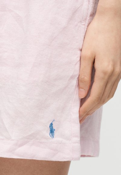 Χέρι που ακουμπά δίπλα σε ανοιχτό ροζ υφασμάτινες σορτς με ένα μικρό μπλε κεντημένο λογότυπο παίκτη πόλο κοντά στο τελείωμα.