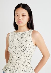 Witte mouwloze top met zwarte stippen en een gegatherde taille; heeft een ronde halslijn en een peplumzoom. Glad, textuurgeweven stof.
