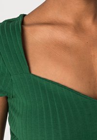 Groene ribbeltop met een V-halsontwerp, voorzien van korte mouwen en een gestructureerde stof. Een gladde huidtoon is zichtbaar bij de halslijn.