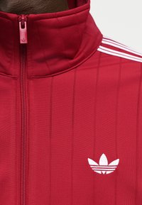 Giacca sportiva rossa con cerniera e collo alto, logo Adidas trefoil bianco sul petto e tre strisce bianche sulla spalla.
